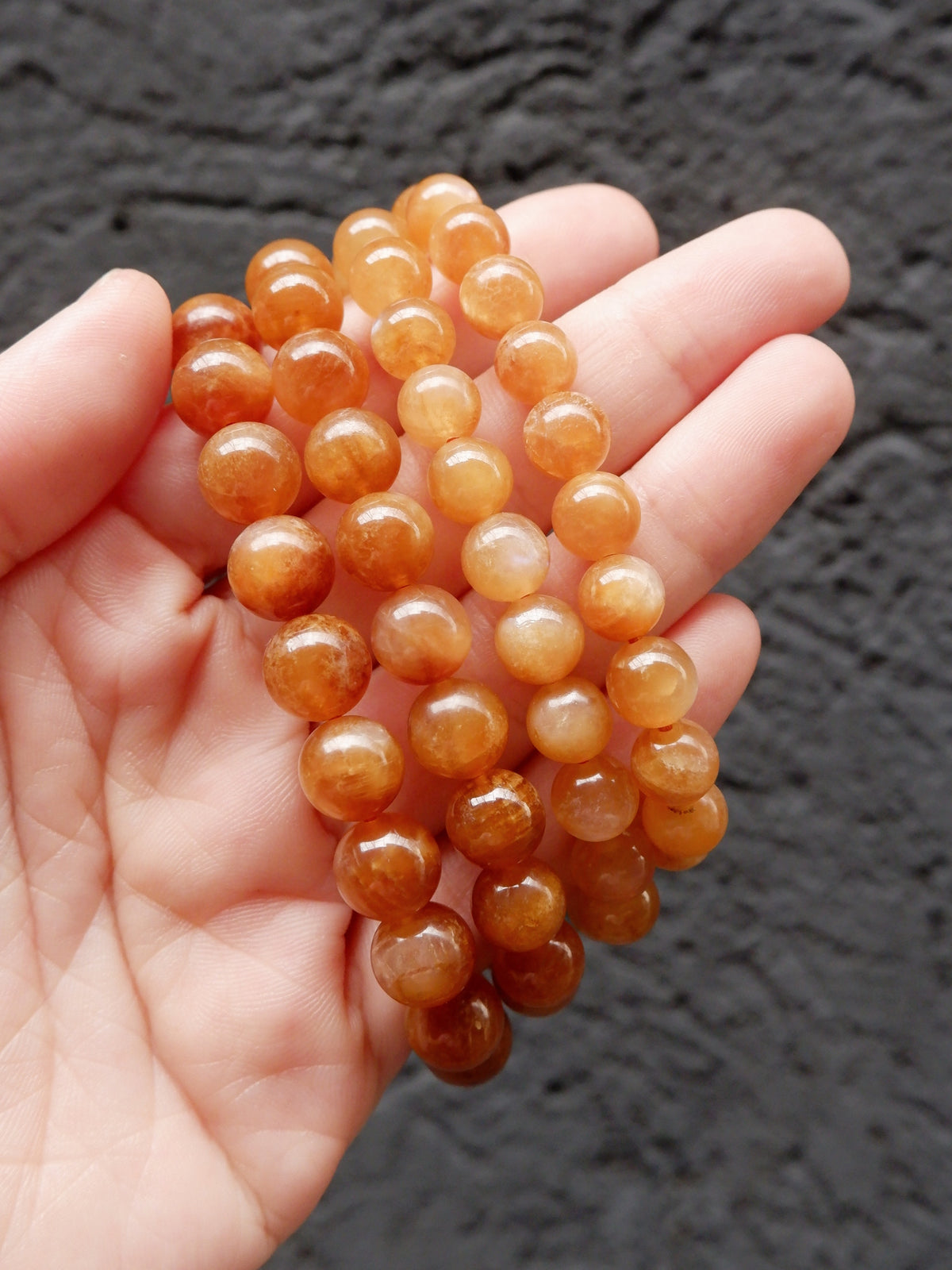 A hand holding three strands of polished orange-brown gemstone beads, likely carnelian, against a dark textured background.