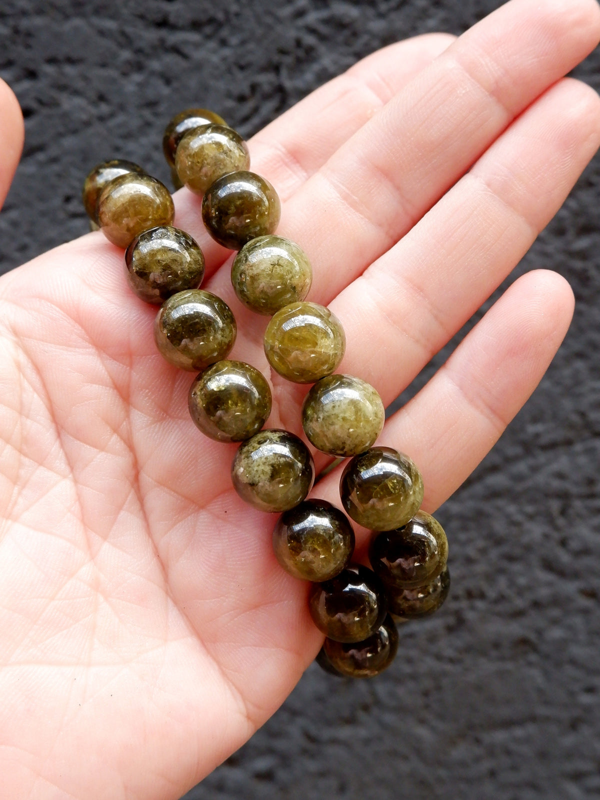 A hand holding a beaded bracelet made of glossy, oval-shaped olive green gemstone beads with natural inclusions, set against a dark textured background.