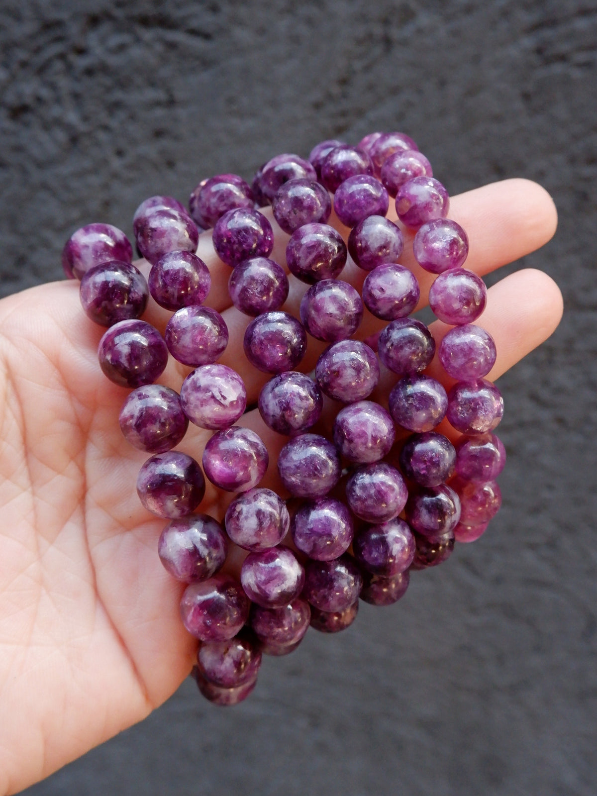 A hand holding a cluster of polished purple gemstone beads, Lepidolite, against a dark textured background.