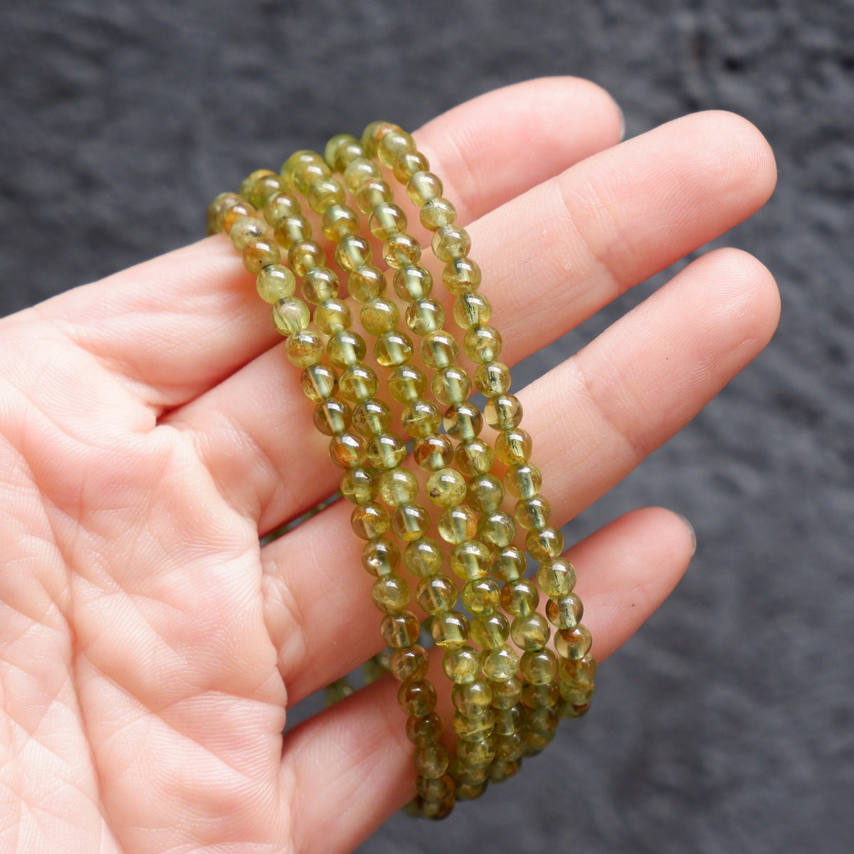 A hand holding a coiled strand of faceted olive green beads against a dark textured background.