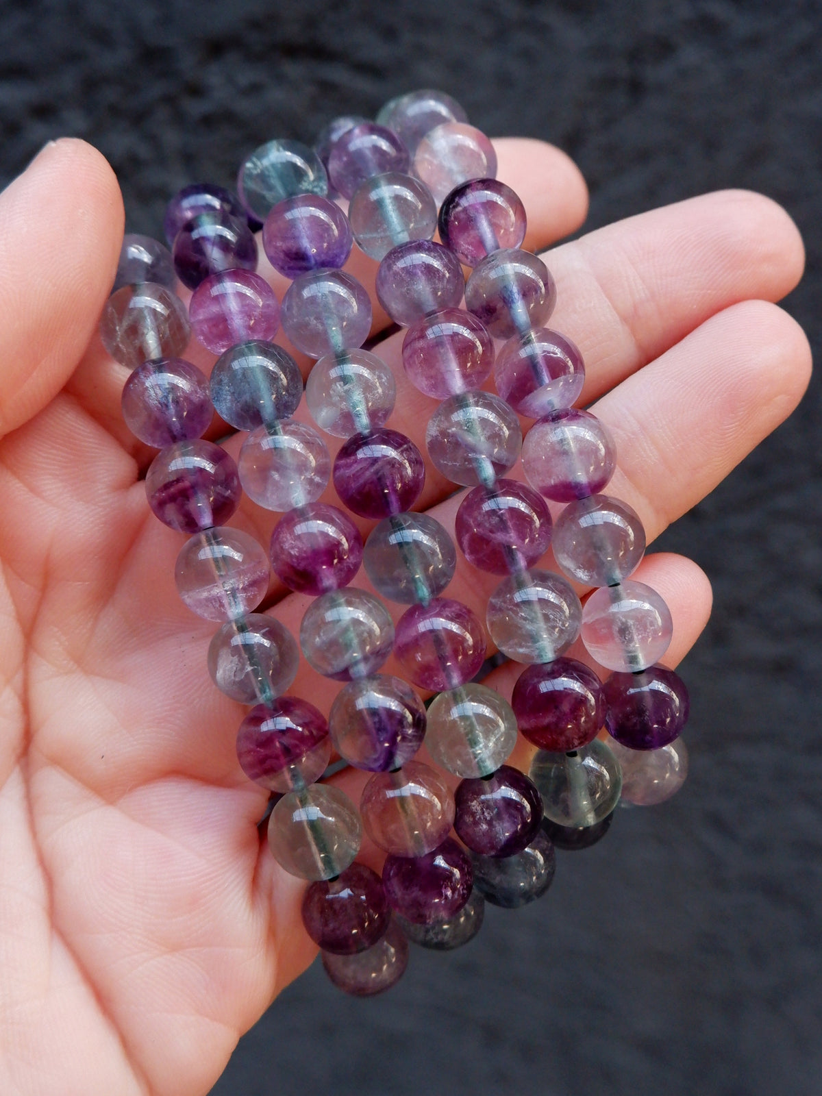 A hand holding a strand of polished round gemstone beads in purple, lavender, and smoky gray tones against a dark textured background.