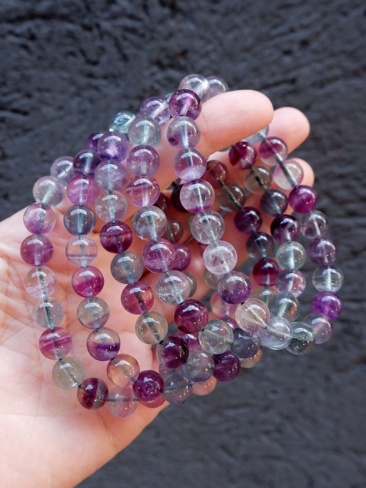 A hand holding a coiled strand of round translucent gemstone beads in shades of purple, lavender, gray, and clear, set against a dark textured background.