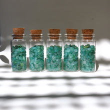 Five small glass bottles with cork lids containing green gemstones on a gray background.