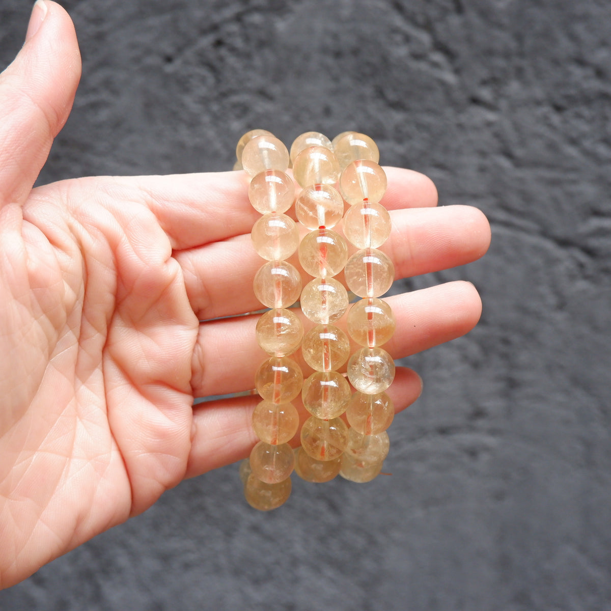 A hand holding three beaded bracelets made of translucent round glass beads in a pale peachy-gold color, set against a dark textured background.