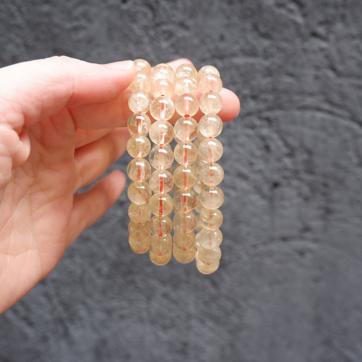 A hand holding four strands of clear round gemstone beads with red thread, shown against a dark textured background.
