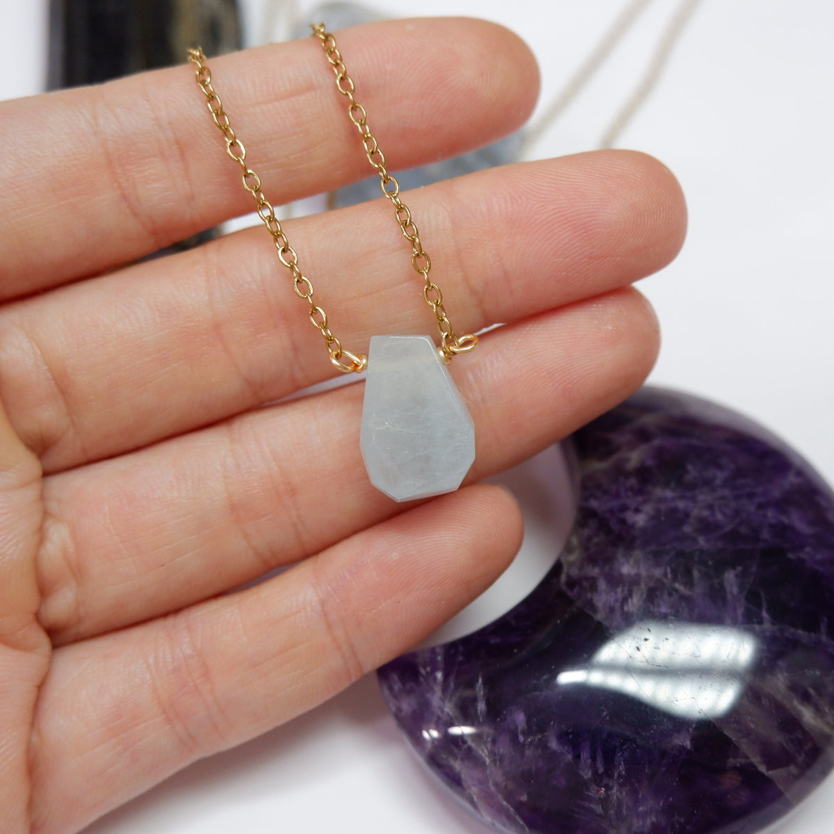 Gold chain necklace with a teardrop-shaped pendant held by a hand, with a purple crystal in the background.