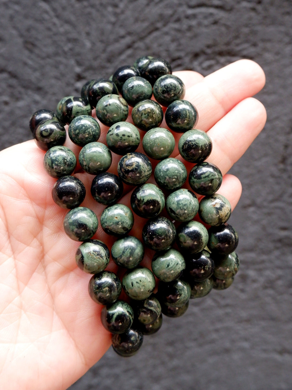 A hand holding a bundle of polished round beads in dark green and black with natural marbled patterns, set against a dark textured background.