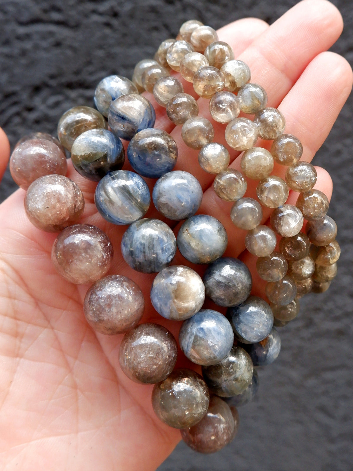 A hand holding multiple strands of polished round gemstone beads, including blue, gray, and smoky brown varieties, against a dark textured background.