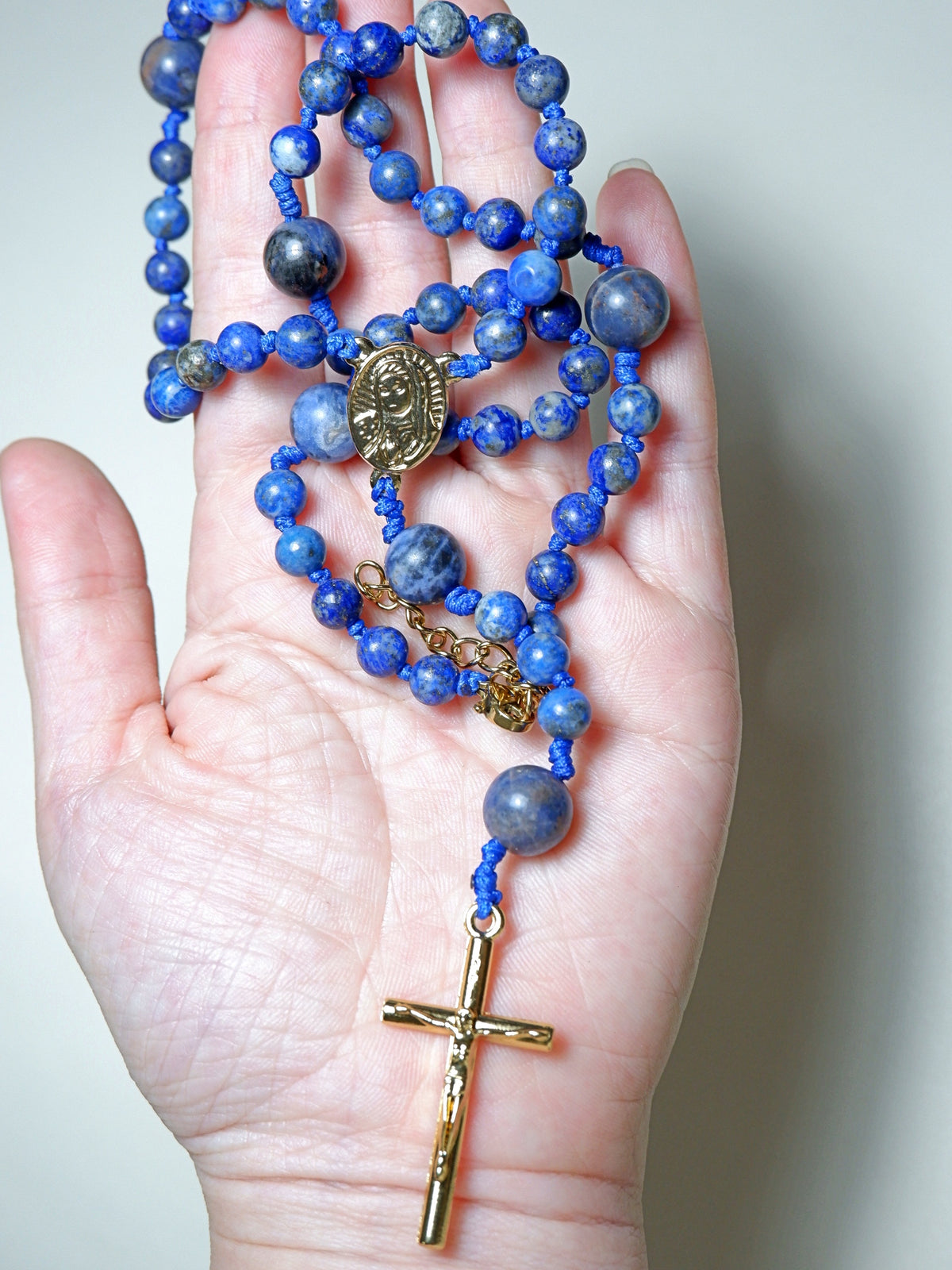 Blue beaded rosary with a gold cross held in a hand against a neutral background