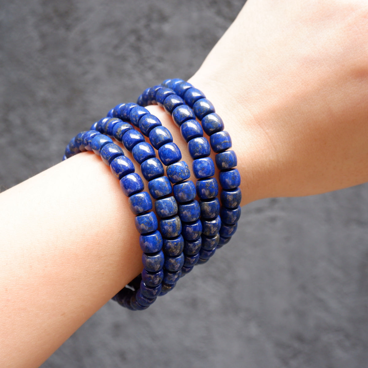 A close-up of a person's wrist wearing five stacked beaded bracelets made of polished deep blue lapis lazuli stones with natural white veining, set against a dark textured gray background.