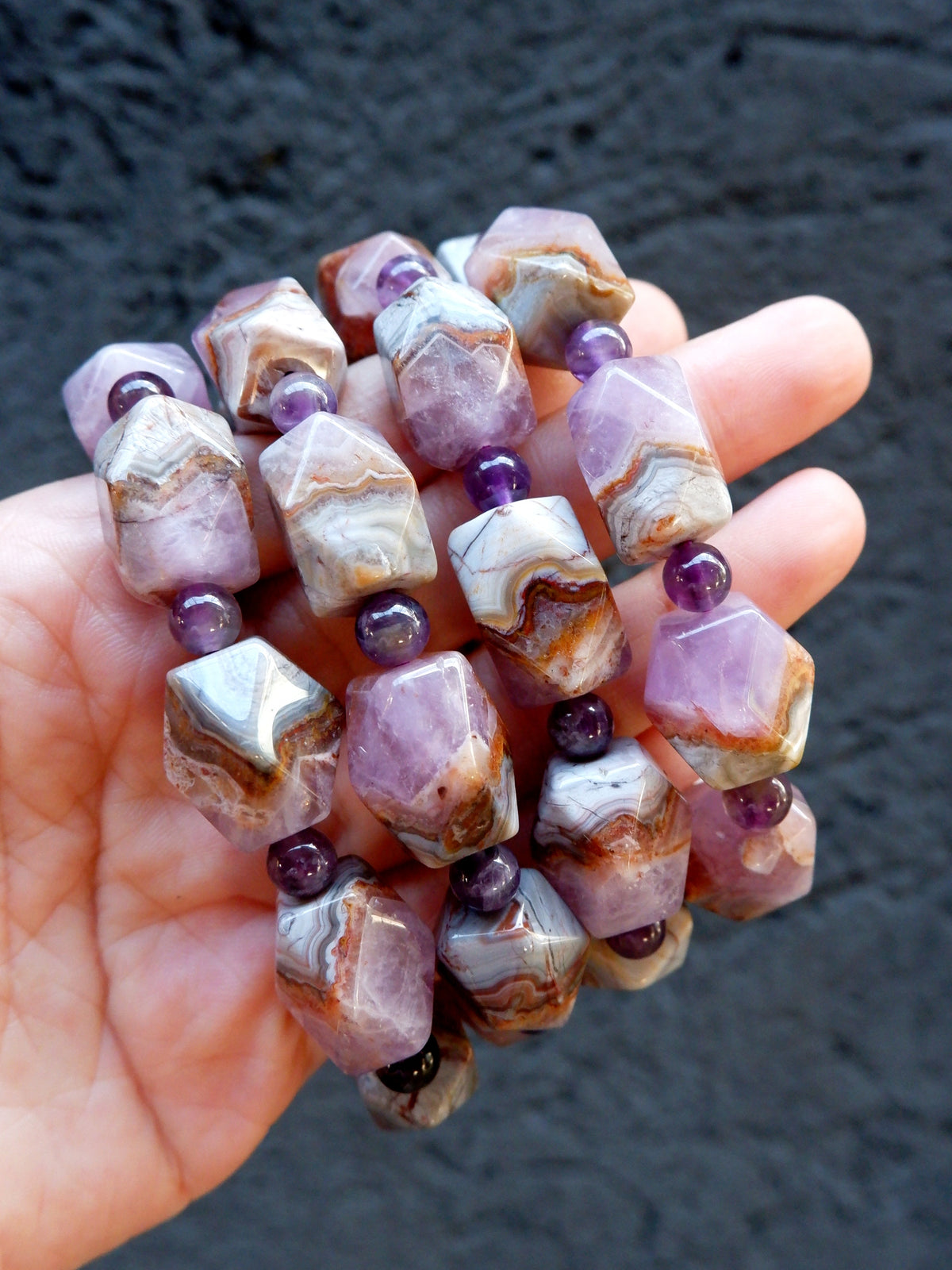 A hand holding a cluster of beaded bracelets made of faceted agate-like beads in lavender, white, and brown bands, separated by small purple beads, against a dark textured background.