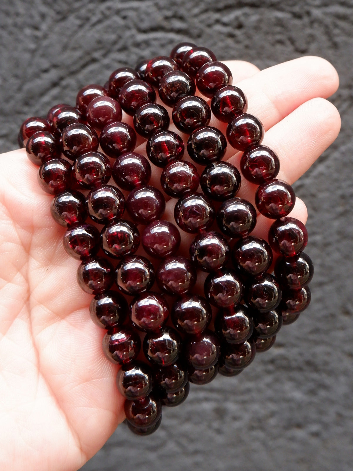 A hand holding a coiled strand of glossy, dark red round beads against a dark textured background.