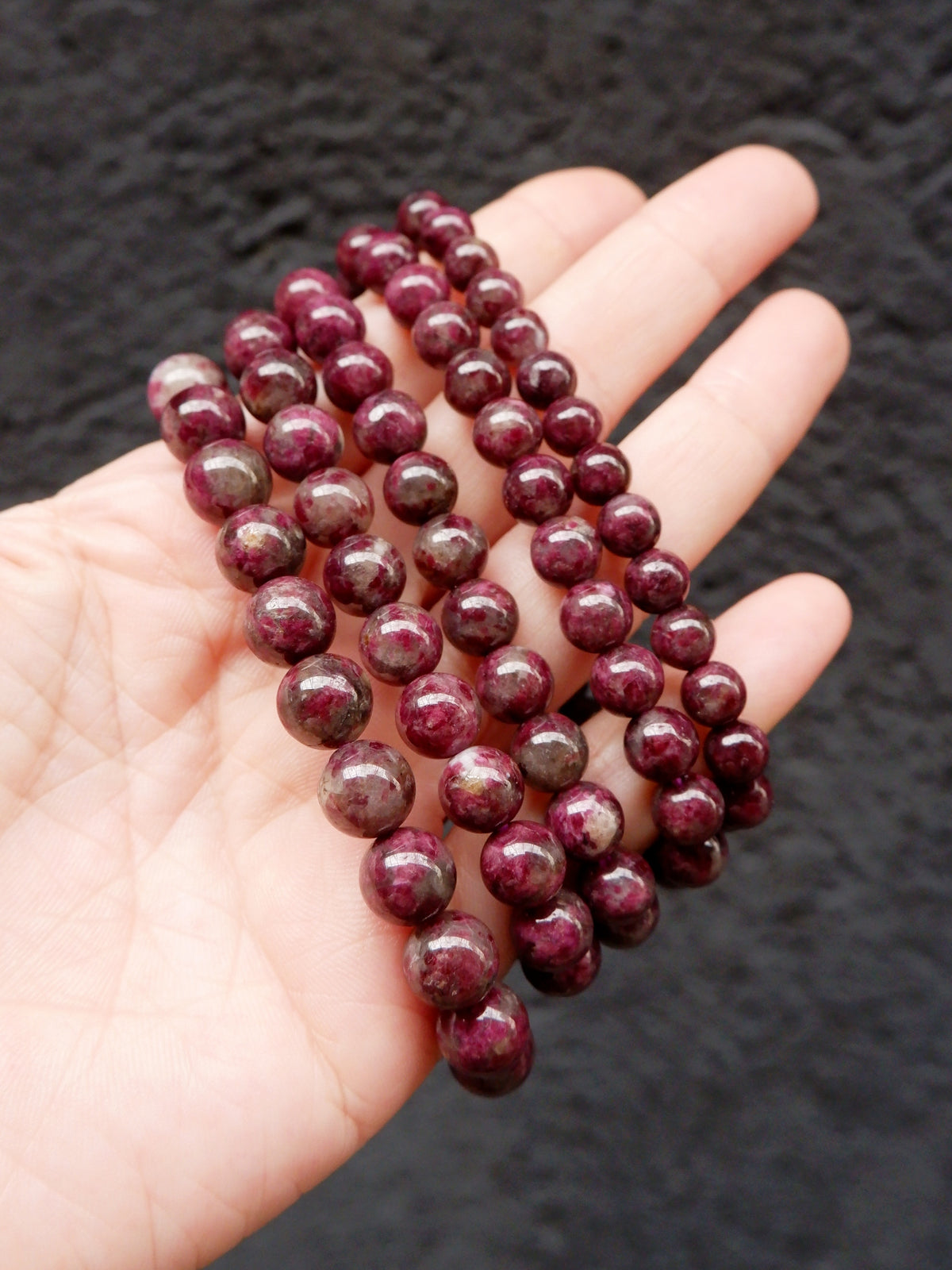 A hand holding three strands of polished dark red tourmaline beads, photographed against a dark textured background.