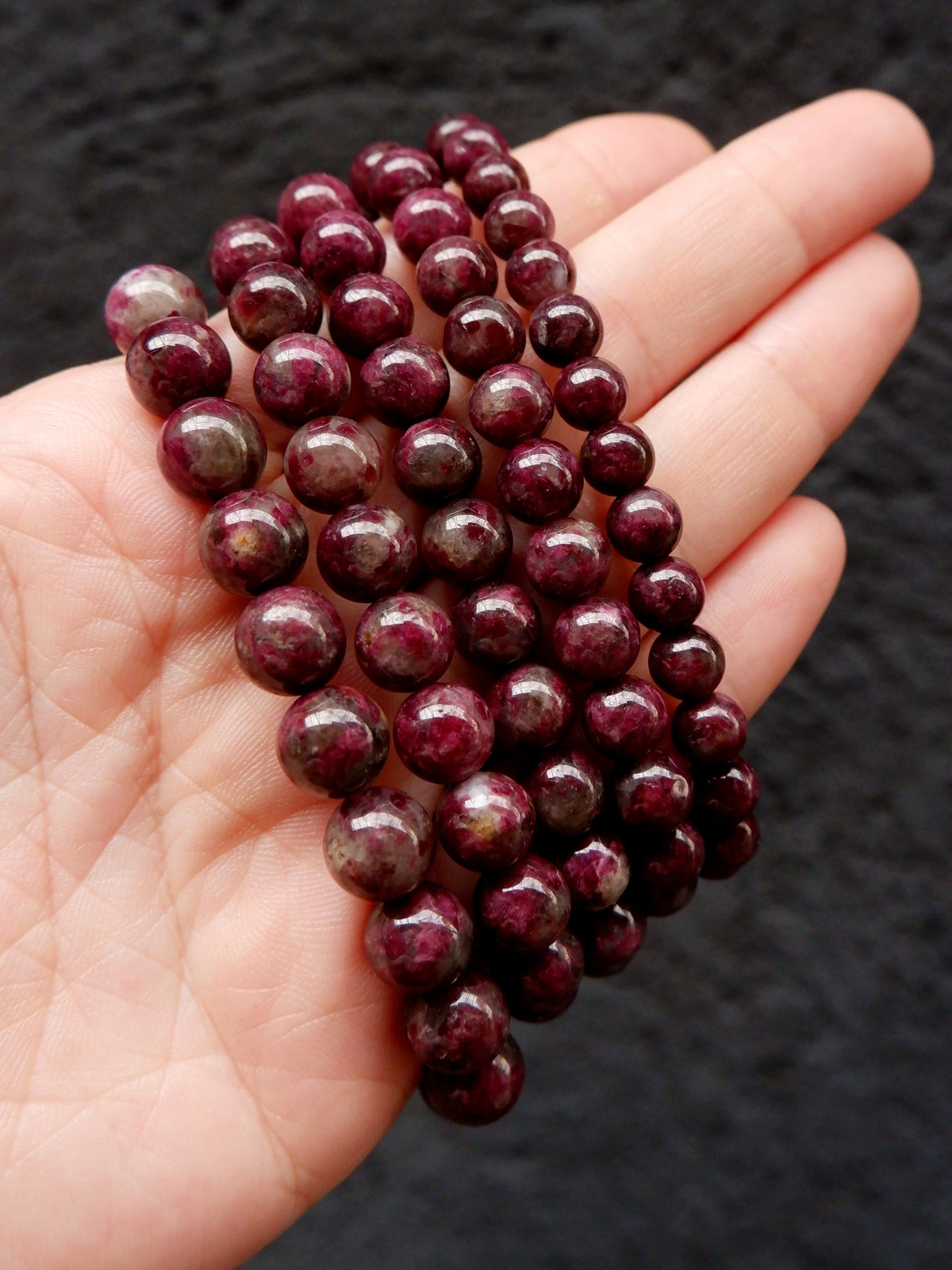 A hand holding a strand of polished round beads in a deep burgundy-purple color, set against a dark textured background.