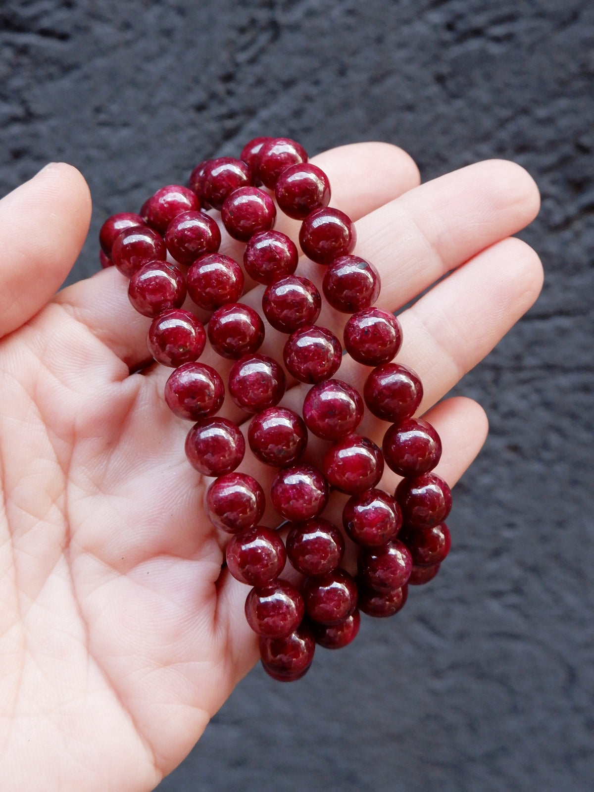 A hand holding a coiled strand of glossy, dark red round beads against a dark textured background.