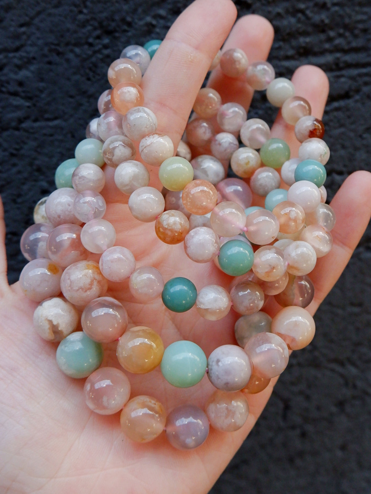 A hand holding a stack of multi-strand beaded bracelets made of polished round gemstone beads in pastel colors including pink, green, blue, orange, and white, set against a dark textured background.