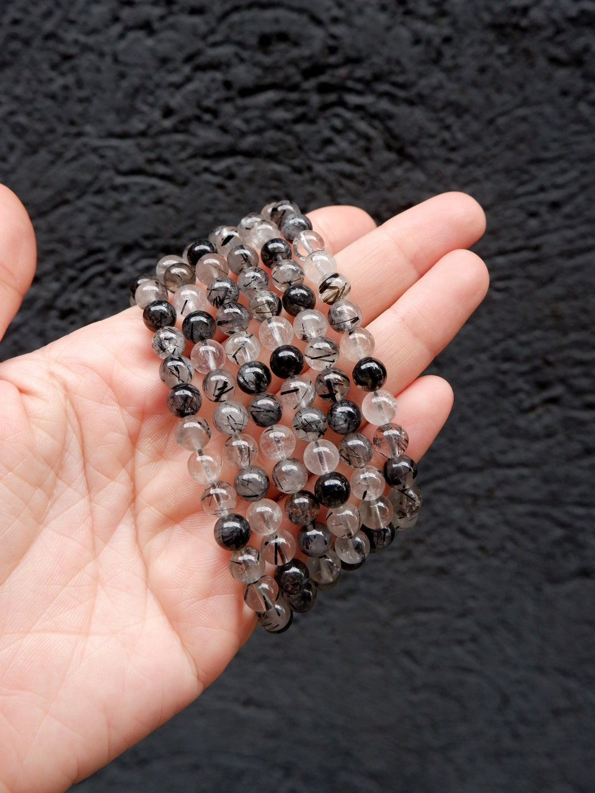 A hand holding a stack of beaded stretch bracelets made of black and smoky gray translucent round beads, set against a dark textured background.