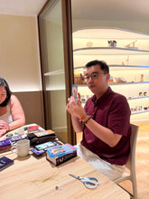 A man in a maroon polo shirt and white pants sits at a wooden table holding a small card or book, surrounded by craft supplies like scissors, a mug, and boxes.