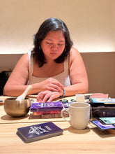 A woman sits at a wooden table with her eyes closed, surrounded by tarot card decks, a brass singing bowl, and a white mug in a warm, indoor setting.