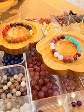Two handmade beaded bracelets displayed on wooden flower-shaped stands, surrounded by clear plastic bins filled with assorted gemstone beads on a wooden table.