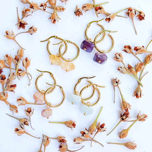 A pair of gold-colored hoop earrings with semi-precious crystals in various colors displayed on a white surface surrounded by dried flower petals.