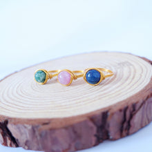 A gold-colored aluminum ring with three different colored semi-precious crystals set in it, placed on a wooden surface.