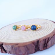A gold-colored aluminum ring with three different colored semi-precious crystals set in it, placed on a wooden surface.