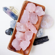 A collection of raw and tumbled rose quartz stones displayed on a wooden plate, surrounded by other crystals.